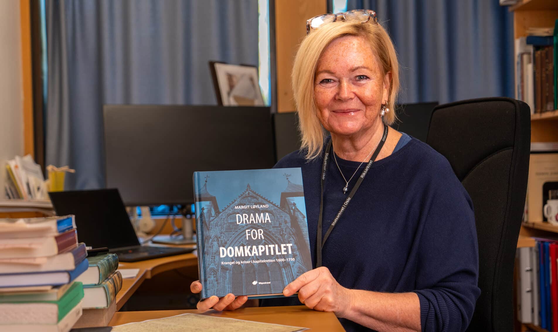Kvinne på kontor holder bok med tittelen Drama for Domkapitlet, omgitt av bokhyller og skrivebord med bærbar PC og papirer.