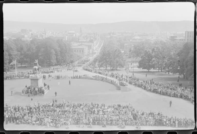 Utsikt fra slottsbalkongen over Slottsplassen og Karl Johans gate. Bildet er tatt i forbindelse med kong Haakons hjemkomst, 7. juni 1945. Fotograf: Nikolai Ramm Østgaard, Arkivref.: Østgaard, Nikolai Ramm og Ragni/Arkivverket/Riksarkivet (RA/PA-1951/U/Ub/L0001).