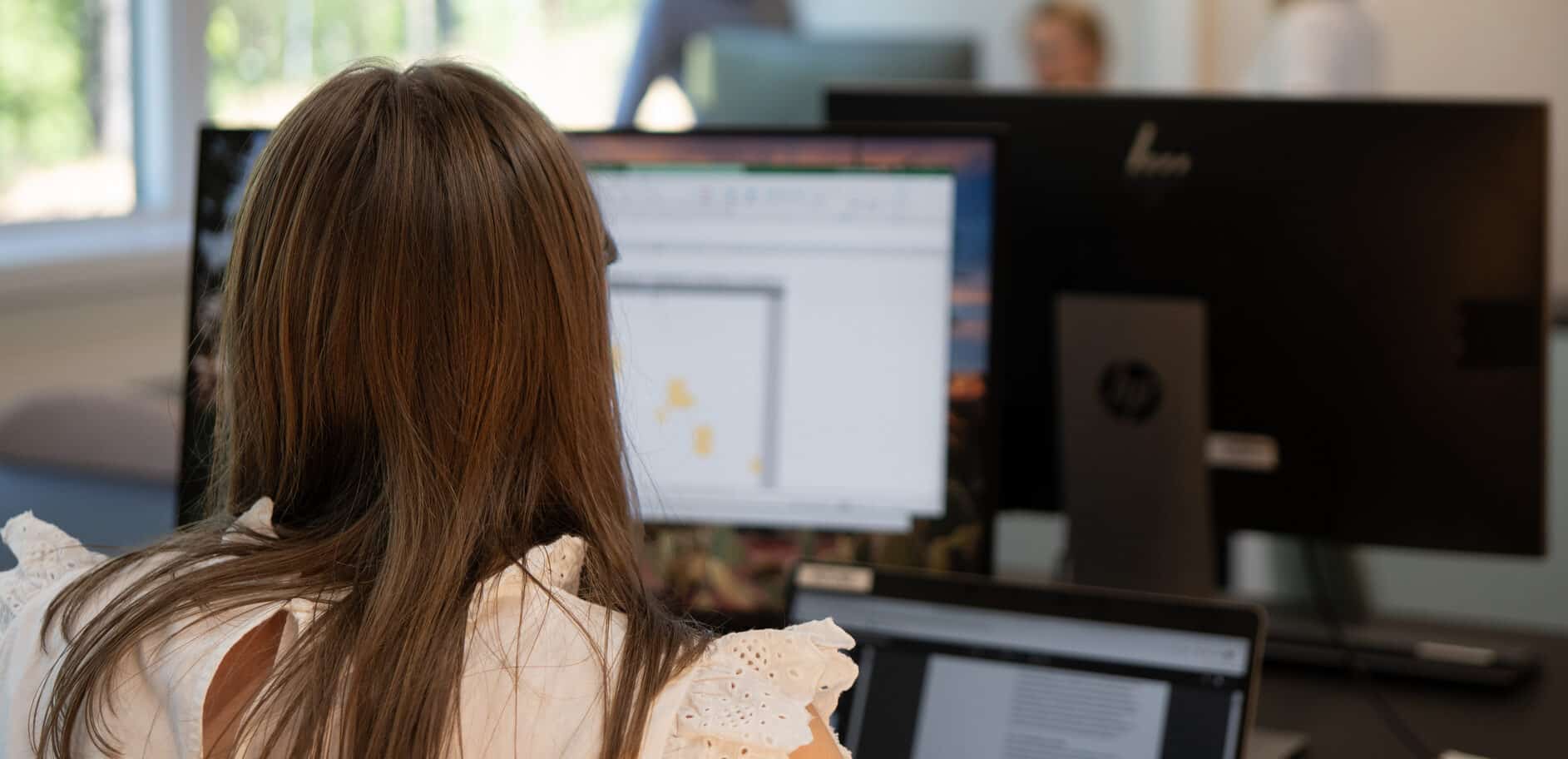 Kvinne som jobber ved datamaskinen på kontoret, kolleger synlige i bakgrunnen. Lys arbeidsplass med vinduer som vender ut mot grøntområder.