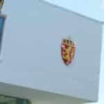 Moderne hvit bygning med norsk våpenskjold, omgitt av trær. Arkitektonisk overheng synlig. Blå himmel i bakgrunnen.