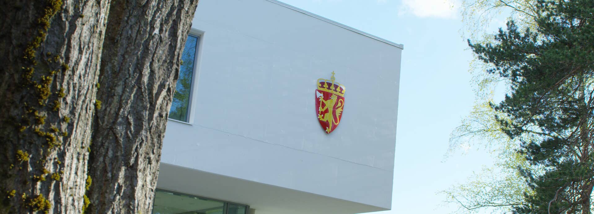 Moderne hvit bygning med norsk våpenskjold, omgitt av trær. Arkitektonisk overheng synlig. Blå himmel i bakgrunnen.