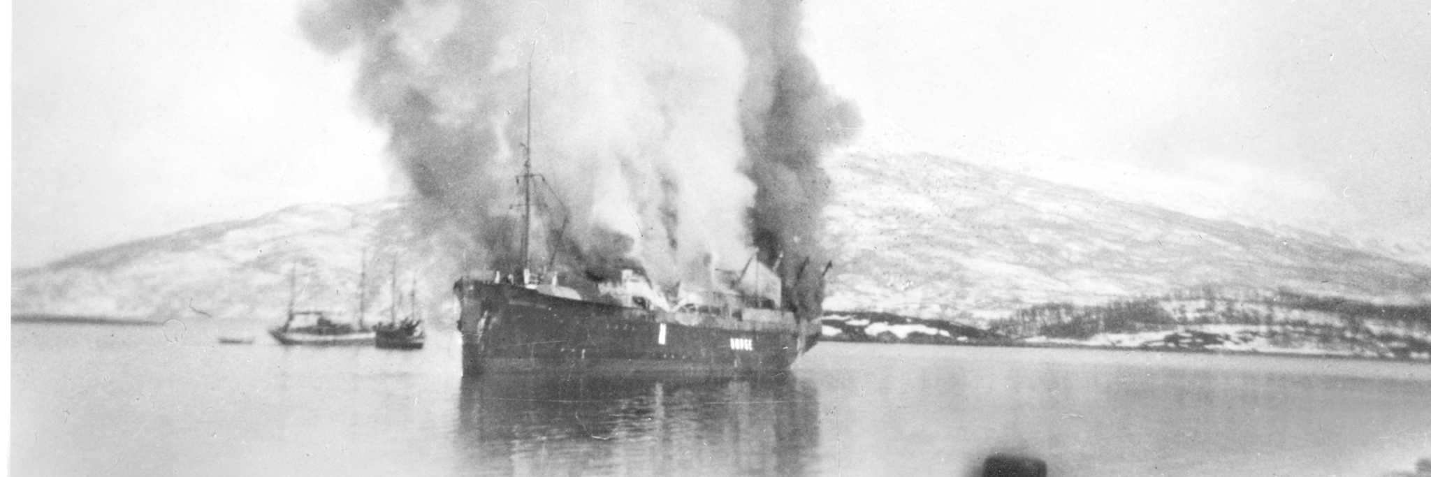 Det tidligere hurtigruteskipet "Dronning Maud" har blitt bombet av tyske fly. Skipet står i brann og mørk røyk velter opp mot himmelen. RA/RAFA-2017/E/Ea/L0088/0013.