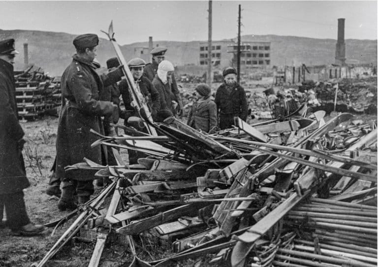 Original bildetekst: "Småbyene og fiskeværene i Nord-Norge ligger som ødeleggelsens monumenter over tysk mordpolitikk, med husene i ruiner og skorsteinspipene stikkende opp mot himlen. I Kirkenes var ødeleggelsen nesten total, bare 28 hus sto tilbake. I Vadsø var 65-70% av byen ødelagt og i Vardø 85%. Innover Varangerfjorden til Vest-Tana er det nesten intet igjen. - Dette lager av ski som sees på bildet rakk tyskerne imidlertid ikke hverken å få ødelagt eller å få med seg. Utstyret studeres av norske offiserer for å bringe på det rene om det kan tas i bruk av troppene". (Arkivreferanse: RA/PA-1209 NTBs krigsarkiv/Digitalarkivet).