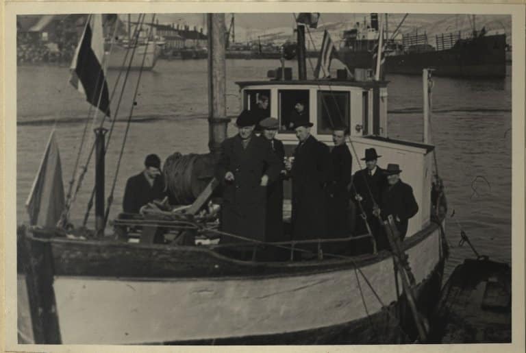 Kirkeskøyta vigsles. Kirkeskøyta på Tromsø havn den 11. april 1946. Ombord sees residerende kapellan Martin Tveter, sokneprest Storvik, sokneprest Notø, Helmer Hansen, biskop Krohn-Hansen og stiftskapellan Amlie. I rorhuset er skipper Peter Solstad og sokneprest Tønder. (SATØ S-1512: Arkivet Biskopen i Nord-Hålogaland, Lpnr. 563, Statsarkivet i Tromsø.)