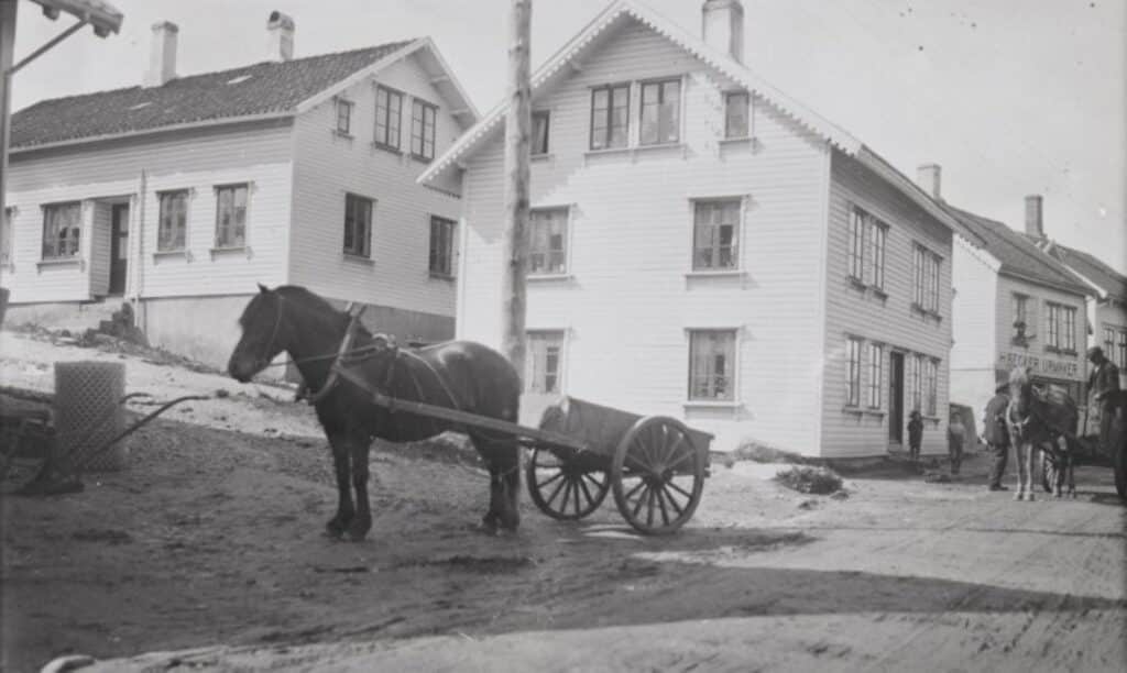 Svart-hvitt-bilde av huset i Storgata 22 på Bryne, med skiltet «H. Becker Urmaker» på fasaden. Et barn står utenfor, og en kvinne skimtes i vinduet. Foto: Peder Netland, 1925.
