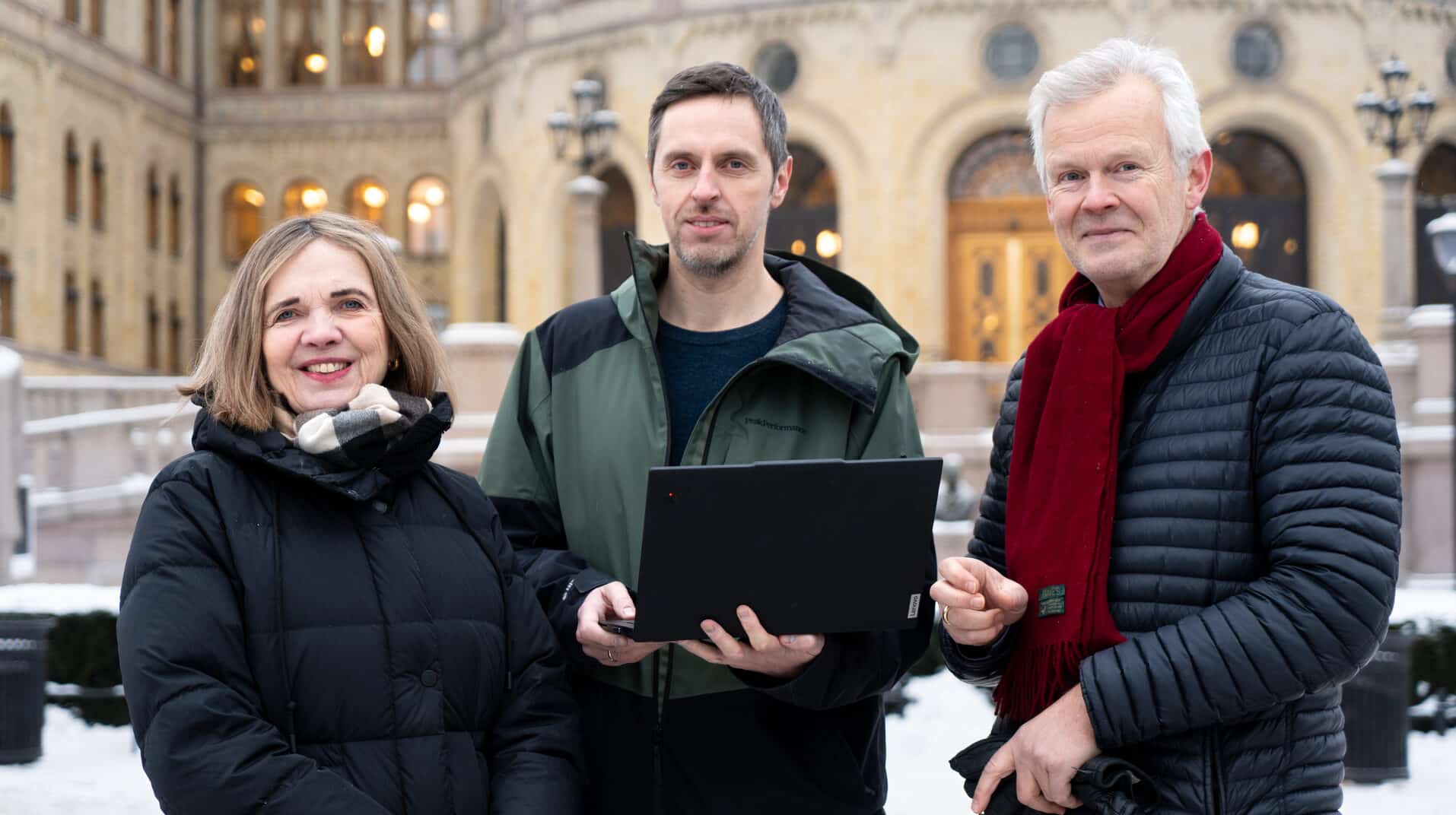 Tre personer står utenfor en snødekt bygning: kvinne i svart frakk, mann med bærbar PC, eldre mann med rødt skjerf.