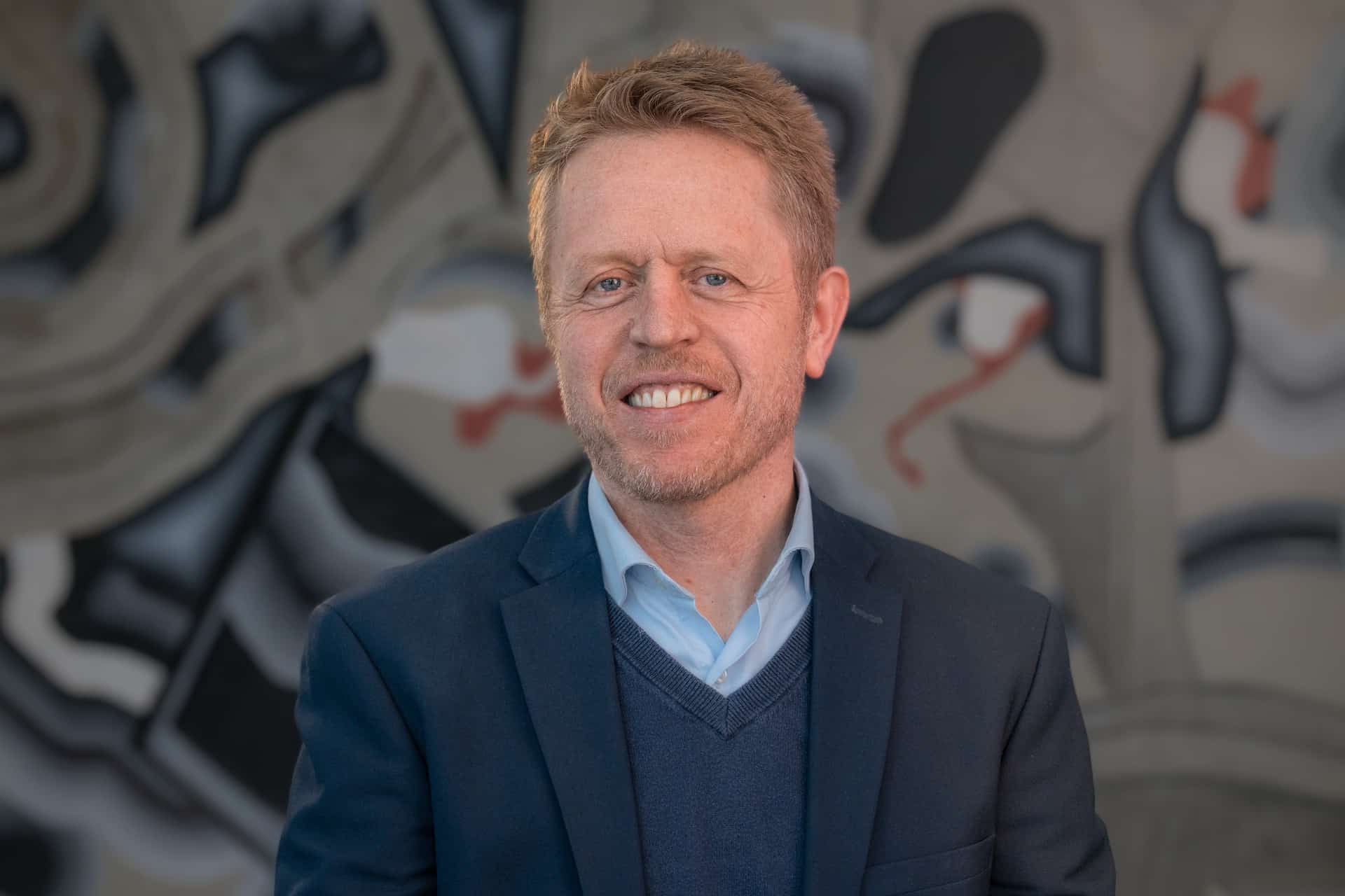 Mann med kort blondt hår smiler til kameraet, iført marineblå dress og lyseblå skjorte mot abstrakt kunstbakgrunn.