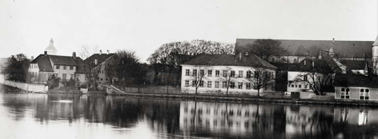 Sort‑hvitt‑fotografi med Breiavatnet i forgrunnen, Alexanders barndomshjem til venstre og Kongsgård skole til høyre.