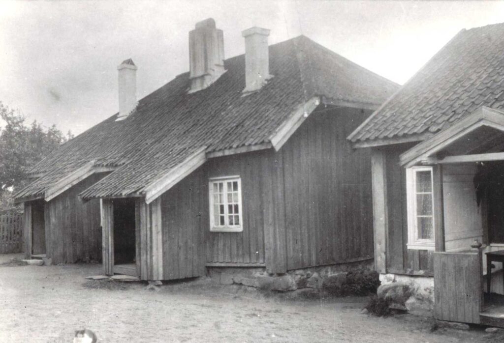 Sort hvitt bilde. Stor arbeiderbolig i tre, med to separate overbygde innganger. Huset har tre piper, en stor på midten og to mindre. Huset er slitent men velholdt. Området rundt er ryddig og rent. En hvit og sort katt ligger på tunet.