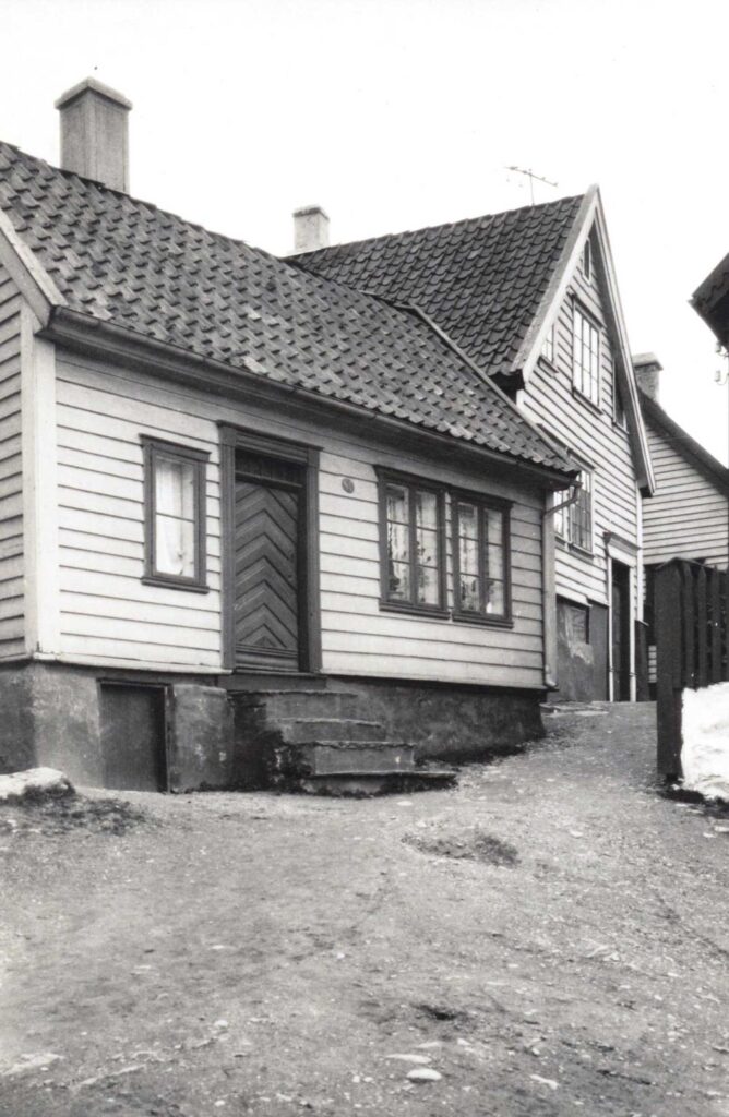 Sort hvitt bilde. Bildet er tatt nede i en smal bratt jordvei. Oppover bildet ser man flere idylliske små trehus. Den nærmeste er hvit med en stentrapp opp til ingangsdøren. Det er et smalt kjøkkenvindu på venstre side av døren og to stuevinduer på den andre siden. Huset fremstår som velholdt og innbydende. En perle mange arbeidere bare kunne drømme om.