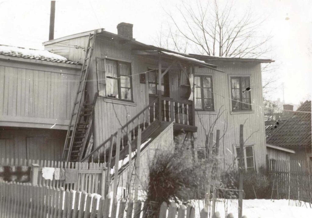 Sort hvitt bilde. Arbeiderbolig i tre med stakittgjerde foran. Det går en trapp på utsiden til en inngangsdør i annen etasje. Huset er ment for i alle fall to familier. Deler av huset virker skjevt men kan være på grunn av kameravinkelen.