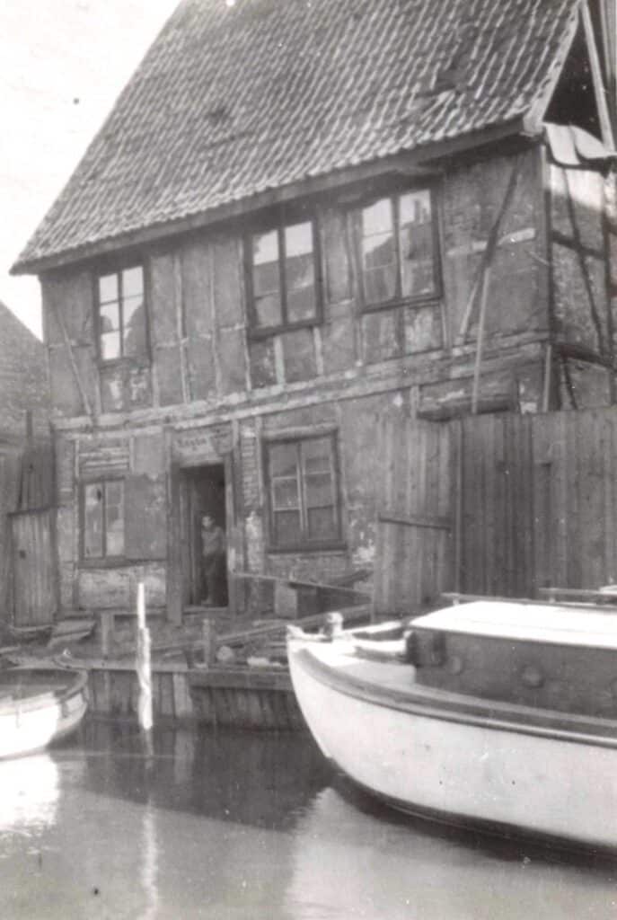 Litt uskarpt sort hvitt bilde. Slitent to etasjers hus rett ved Akerselva. Huset er i binningsverk med synlig tømmer og murstein. Huset og taket er skjevt. I døråpningen står en gutt å ser bort på fotografen. Det ligger to små båter fortøyd foran huset.
