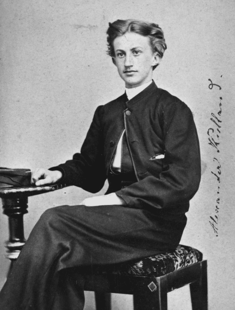 Sort‑hvitt portrett av en ung Alexander L. Kielland, sittende på en krakk med én hånd hvilende på et lite bord. Bildet viser ham i helfigur, eller nesten helfigur.