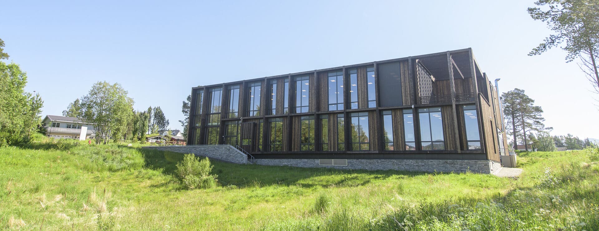 Moderne toetasjes bygning med glassfasade på en gresskledd bakke, omgitt av trær under klar blå himmel.