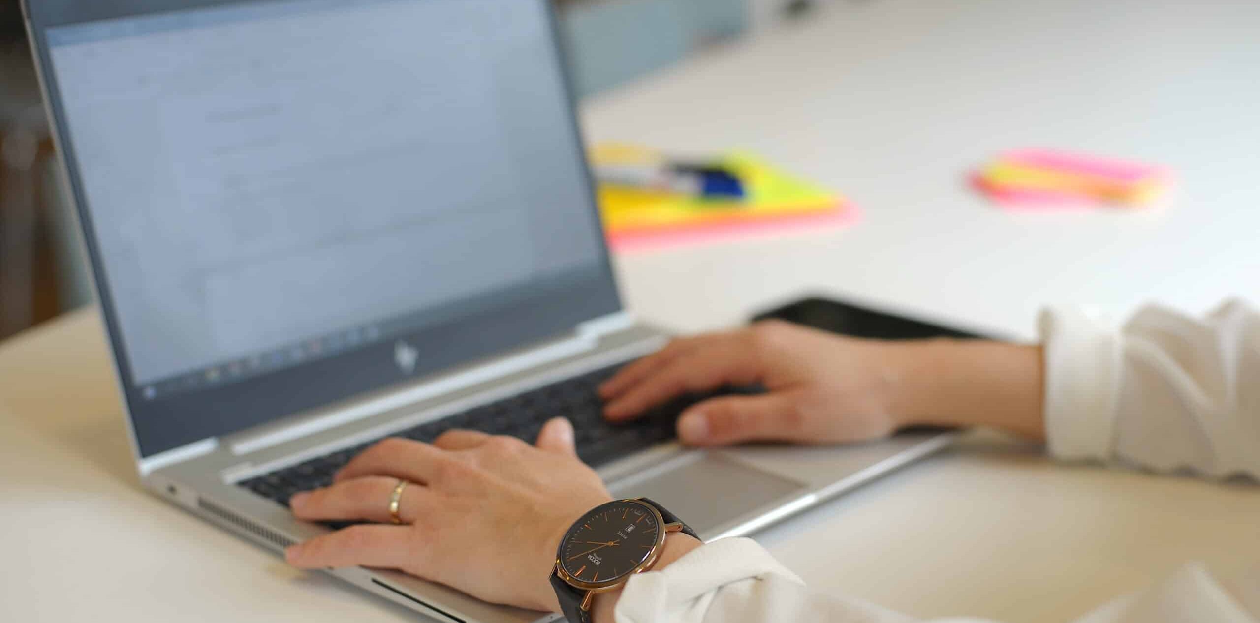 Person som skriver på bærbar PC, iført hvit skjorte og armbåndsur. Fargerike klistrelapper synlige på skrivebordet. Fokus på hender og datamaskin.