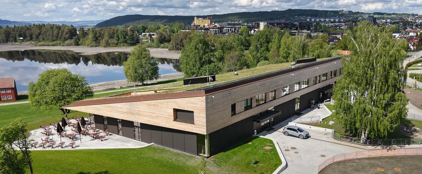 Moderne bygg med tre- og glassfasade sett ovenfra, omgitt av grøntområde og med utsikt over et vann. I bakgrunnen sees skogkledde åser og bebyggelse under en skyet himmel.