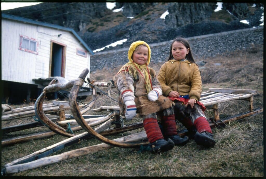 To samiske barn sitter på en tradisjonell treslede utenfor et hus i et fjellandskap. Det ene barnet har kofte og pelsklær, mens det andre har en brun jakke. I bakgrunnen ligger steinete fjell med snøflekker.