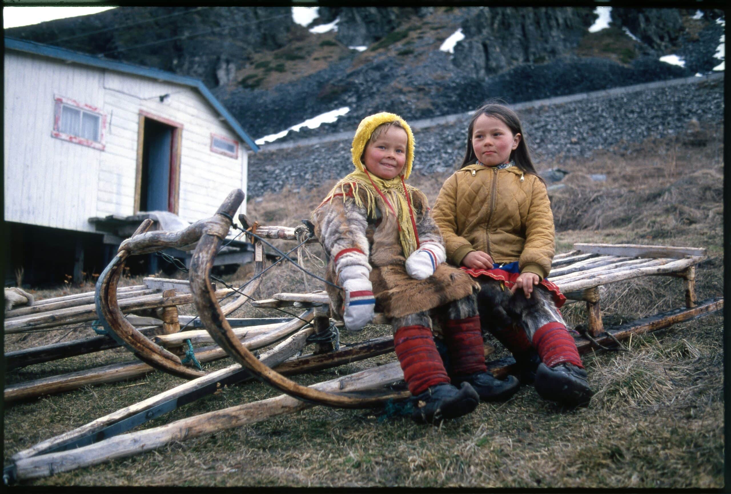 To samiske barn sitter på en tradisjonell treslede utenfor et hus i et fjellandskap. Det ene barnet har kofte og pelsklær, mens det andre har en brun jakke. I bakgrunnen ligger steinete fjell med snøflekker.