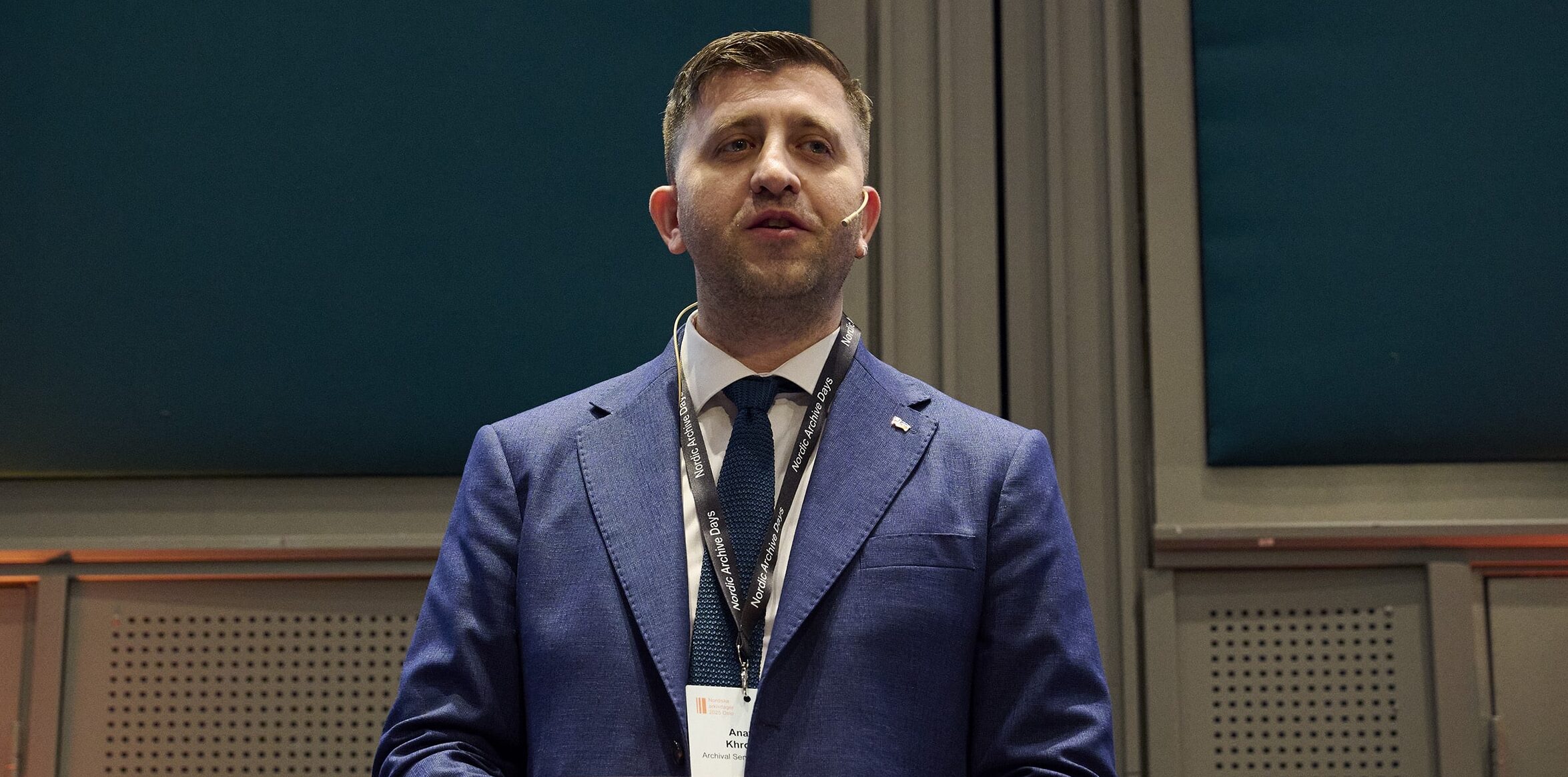 Mann i blå dress på podiet med konferansemerke, snakker med mikrofon i et profesjonelt miljø.