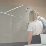 Gruppe mennesker på kontor undersøker whiteboard med bærbar PC på bordet. Moderne arbeidsplass med glassvegger og minimalistiske møbler.