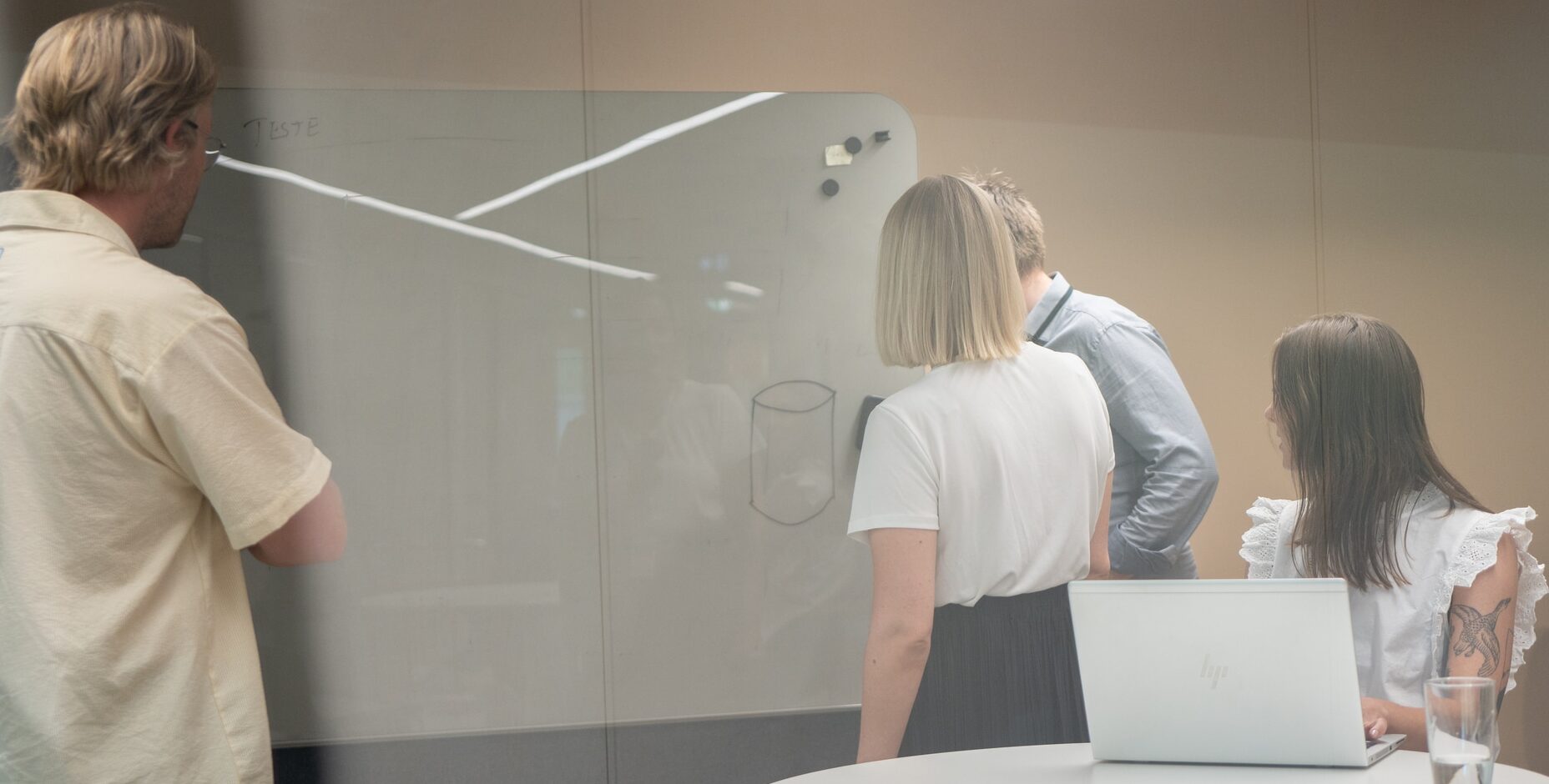Gruppe mennesker på kontor undersøker whiteboard med bærbar PC på bordet. Moderne arbeidsplass med glassvegger og minimalistiske møbler.