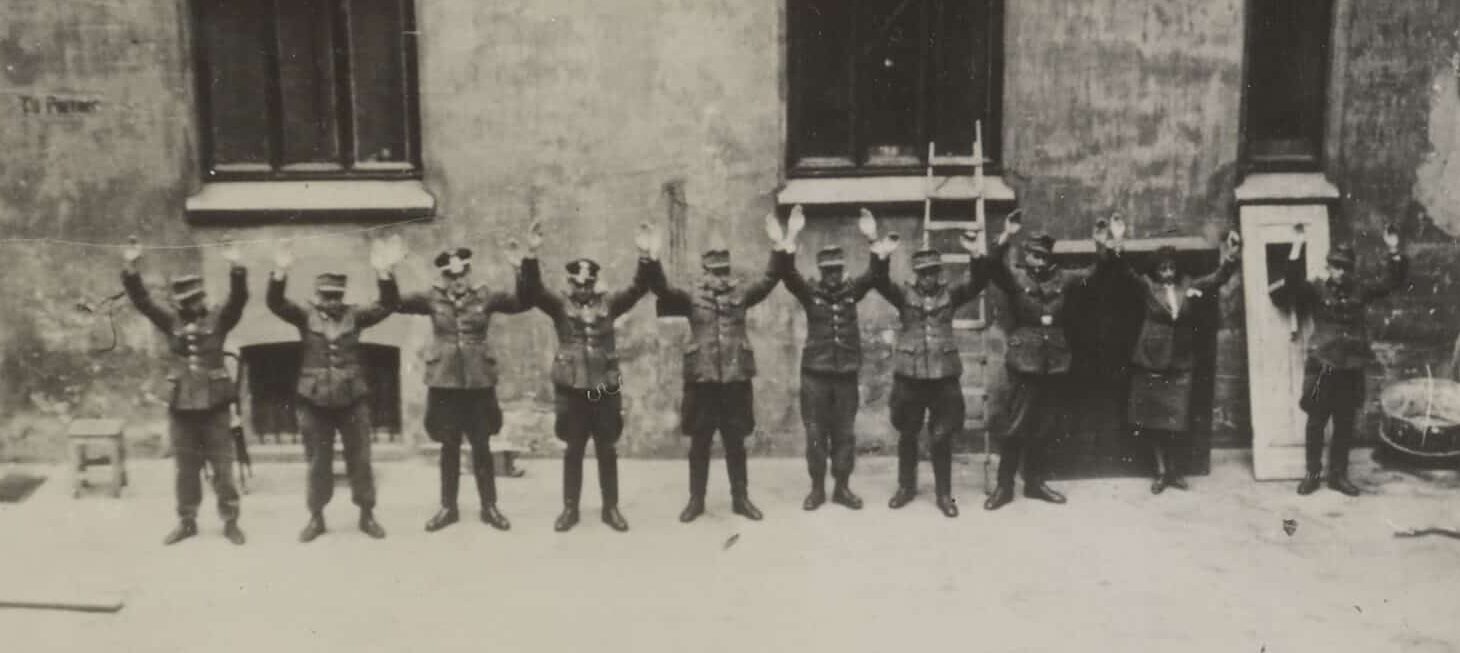 Svart-hvitt-fotografi av en gruppe uniformerte menn som står på rekke foran en bygning. De holder armene løftet over hodet.
