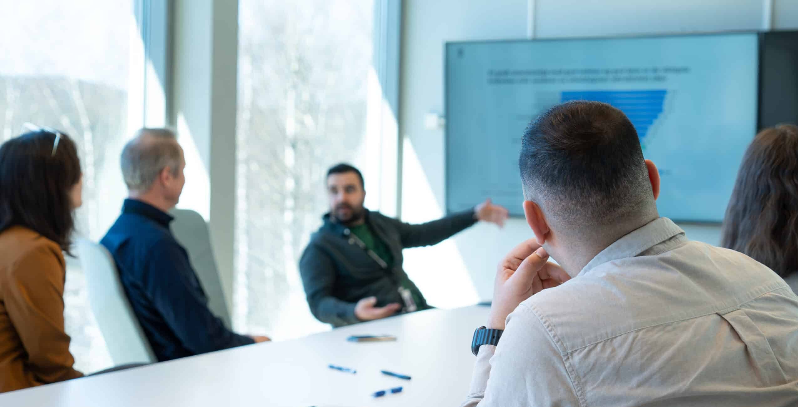 En gruppe mennesker i et møterom som oppmerksomt ser på en presentasjon på en skjerm.