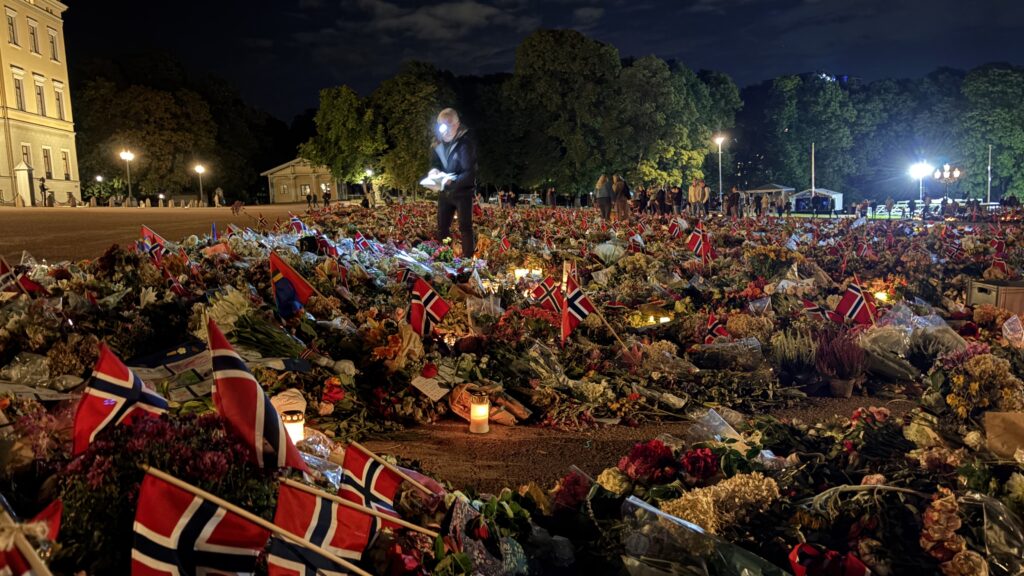 Et stort hav av blomster, norske flagg, lys og hilsener utenfor Slottet i Oslo om kvelden. Mennesker beveger seg mellom minnegjenstandene.